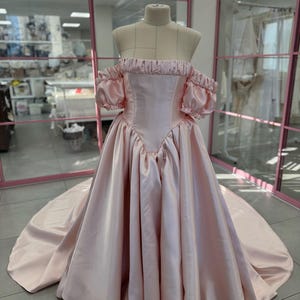 Può includere: Un abito rosa chiaro con spalle scoperte su un manichino. L'abito presenta un corpetto aderente, maniche a sbuffo e una gonna ampia con un lungo strascico. Il tessuto satinato è lucido e l'abito è esposto in una stanza luminosa.