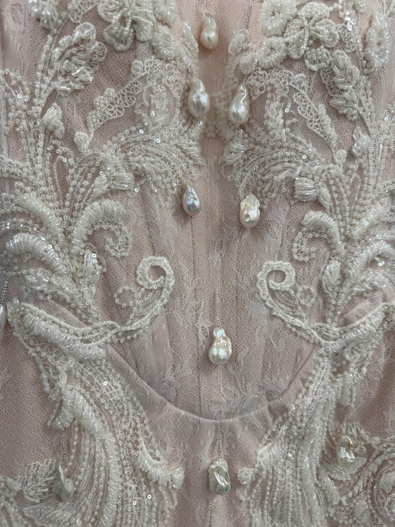 Pu&ograve; includere: Un corpetto in pizzo rosa pallido con intricati ricami floreali bianchi, paillettes trasparenti e perle pendenti a forma di goccia. Il capo presenta un design strutturato con delicati strati di pizzo e una serie di perle applicate.