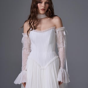 May include: A white floor-length gown featuring a pleated bodice and a sweetheart neckline. The dress includes detachable long sheer sleeves with ruffled cuffs and a matching sheer choker collar, all crafted from a delicate, vertically pleated fabric.