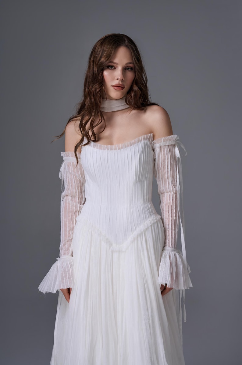 May include: A white floor-length gown featuring a pleated bodice and a sweetheart neckline. The dress includes detachable long sheer sleeves with ruffled cuffs and a matching sheer choker collar, all crafted from a delicate, vertically pleated fabric.