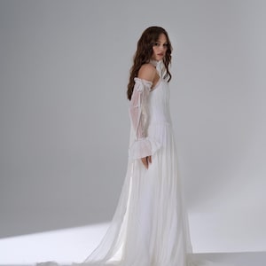 May include: A floor-length white gown featuring a pleated bodice, a high neckline, and sheer off-the-shoulder sleeves with ruffled cuffs. The skirt is made of flowing, layered fabric that extends into a long train, creating a soft silhouette.