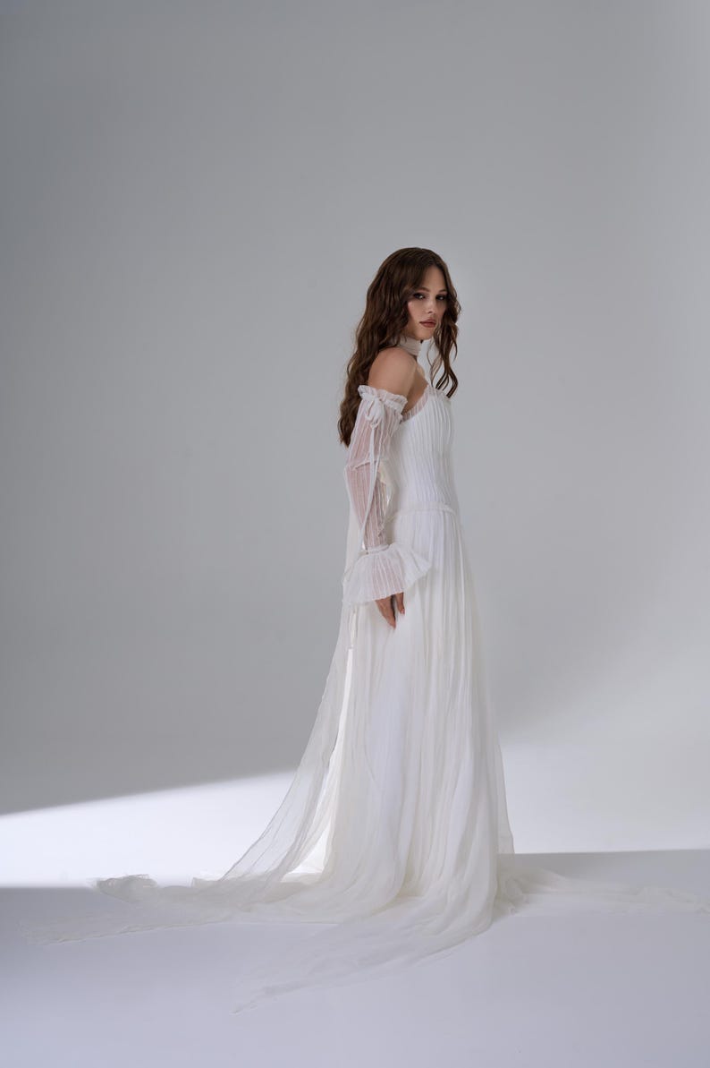 May include: A floor-length white gown featuring a pleated bodice, a high neckline, and sheer off-the-shoulder sleeves with ruffled cuffs. The skirt is made of flowing, layered fabric that extends into a long train, creating a soft silhouette.
