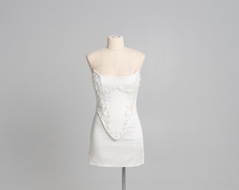 Peut inclure: Une robe courte blanche sans bretelles de style corset avec un ourlet en pointe et une broderie florale complexe ornée de petites perles blanches sur le corsage et les côtés, présentée sur un mannequin sur fond gris clair.