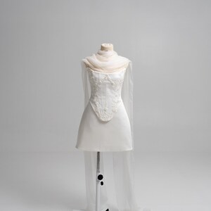 Puede incluir: Un vestido corto blanco sin tirantes con un corpi&ntilde;o estructurado que presenta intrincados bordados florales blancos y pedrer&iacute;a. La prenda incluye un chal beige p&aacute;lido, ligero y transparente, drapeado alrededor del cuello y con paneles largos.