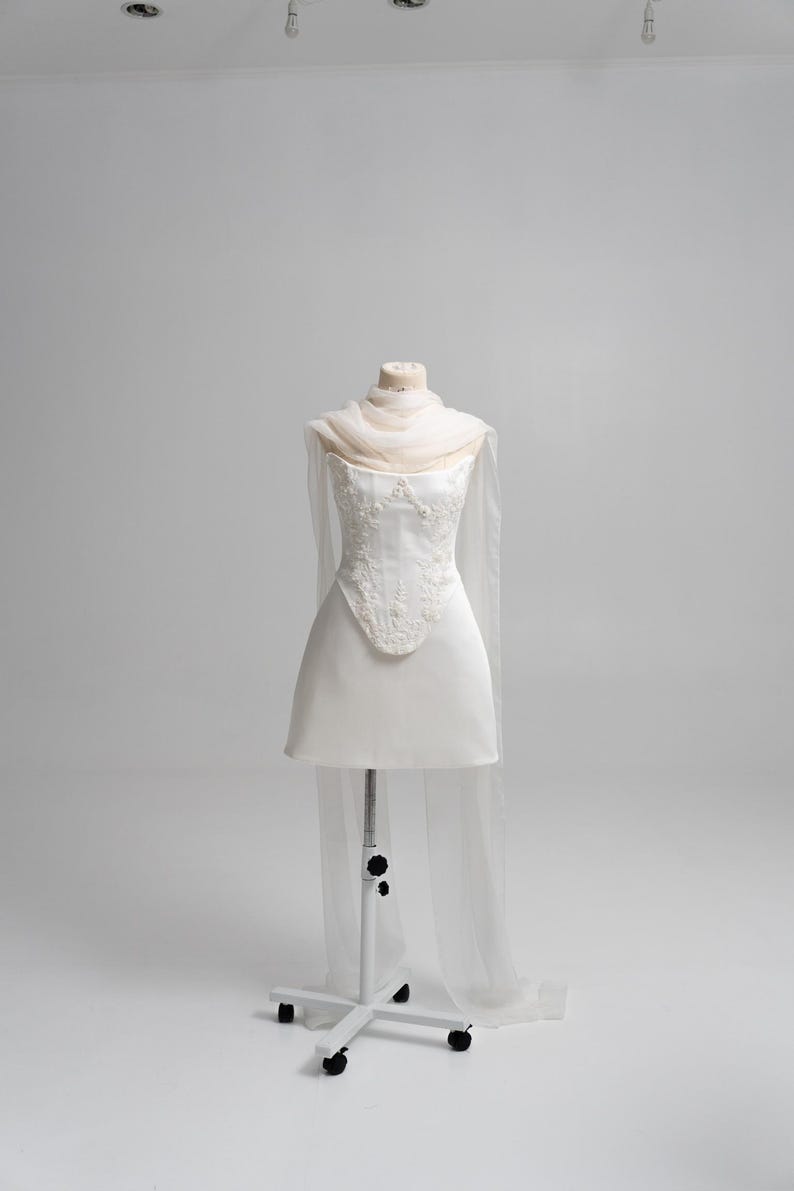 Puede incluir: Un vestido corto blanco sin tirantes con un corpi&ntilde;o estructurado que presenta intrincados bordados florales blancos y pedrer&iacute;a. La prenda incluye un chal beige p&aacute;lido, ligero y transparente, drapeado alrededor del cuello y con paneles largos.