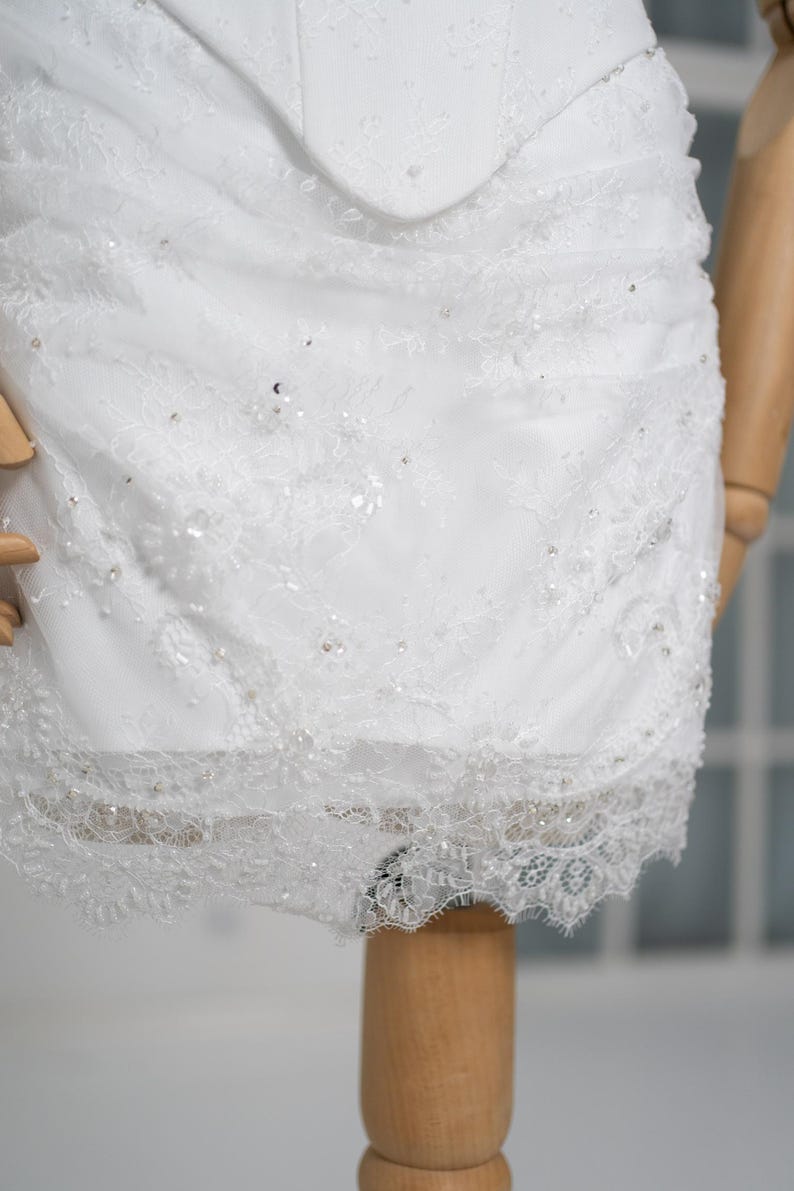 May include: Close-up of a white wedding dress bodice and skirt. The dress features intricate lace detailing, delicate beading, and a scalloped lace hem. The dress is displayed on a wooden mannequin.