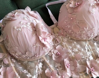 May include: A light pink floral corset and bra set. The bra cups and corset are adorned with delicate, three-dimensional flowers and lace detailing. The set features adjustable straps and a structured design, perfect for special occasions.