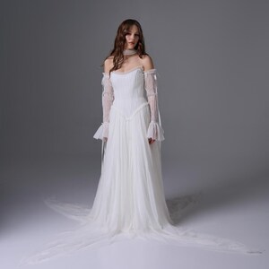 May include: A floor-length white gown featuring a pleated bodice, an off-the-shoulder neckline, and long, sheer, ruffled sleeves. The skirt is made of layered tulle, creating a flowing silhouette that extends into a train on the floor.