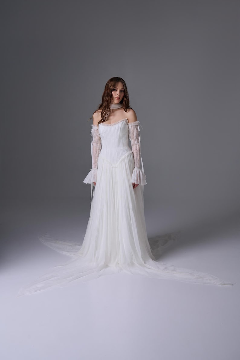 May include: A floor-length white gown featuring a pleated bodice, an off-the-shoulder neckline, and long, sheer, ruffled sleeves. The skirt is made of layered tulle, creating a flowing silhouette that extends into a train on the floor.
