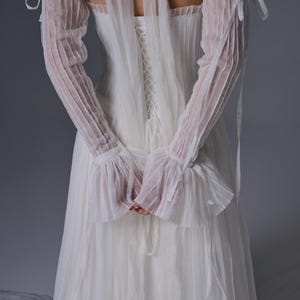 May include: A white floor-length dress featuring a corset-style back with lace-up detailing, paired with matching sheer, pleated, off-the-shoulder sleeves that tie at the upper arms and feature ruffled cuffs, all crafted from a lightweight fabric.