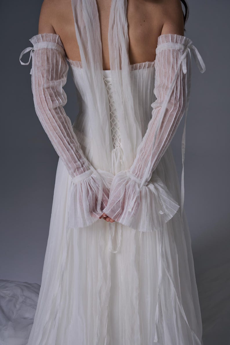 May include: A white floor-length dress featuring a corset-style back with lace-up detailing, paired with matching sheer, pleated, off-the-shoulder sleeves that tie at the upper arms and feature ruffled cuffs, all crafted from a lightweight fabric.