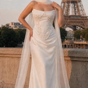 Puede incluir: Un vestido largo blanco sin tirantes con un corpi&ntilde;o estructurado que presenta intrincados bordados florales y pedrer&iacute;a. El vestido tiene una falda drapeada y un chal largo de tela ligera y transparente que cae desde los hombros al suelo.