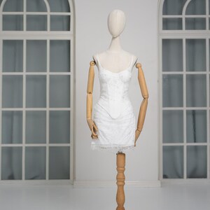 May include: A white, lace-detailed mini dress with spaghetti straps is displayed on a wooden mannequin. The dress features a fitted bodice and a flared skirt, with a delicate lace trim at the hem. The mannequin has articulated wooden arms.