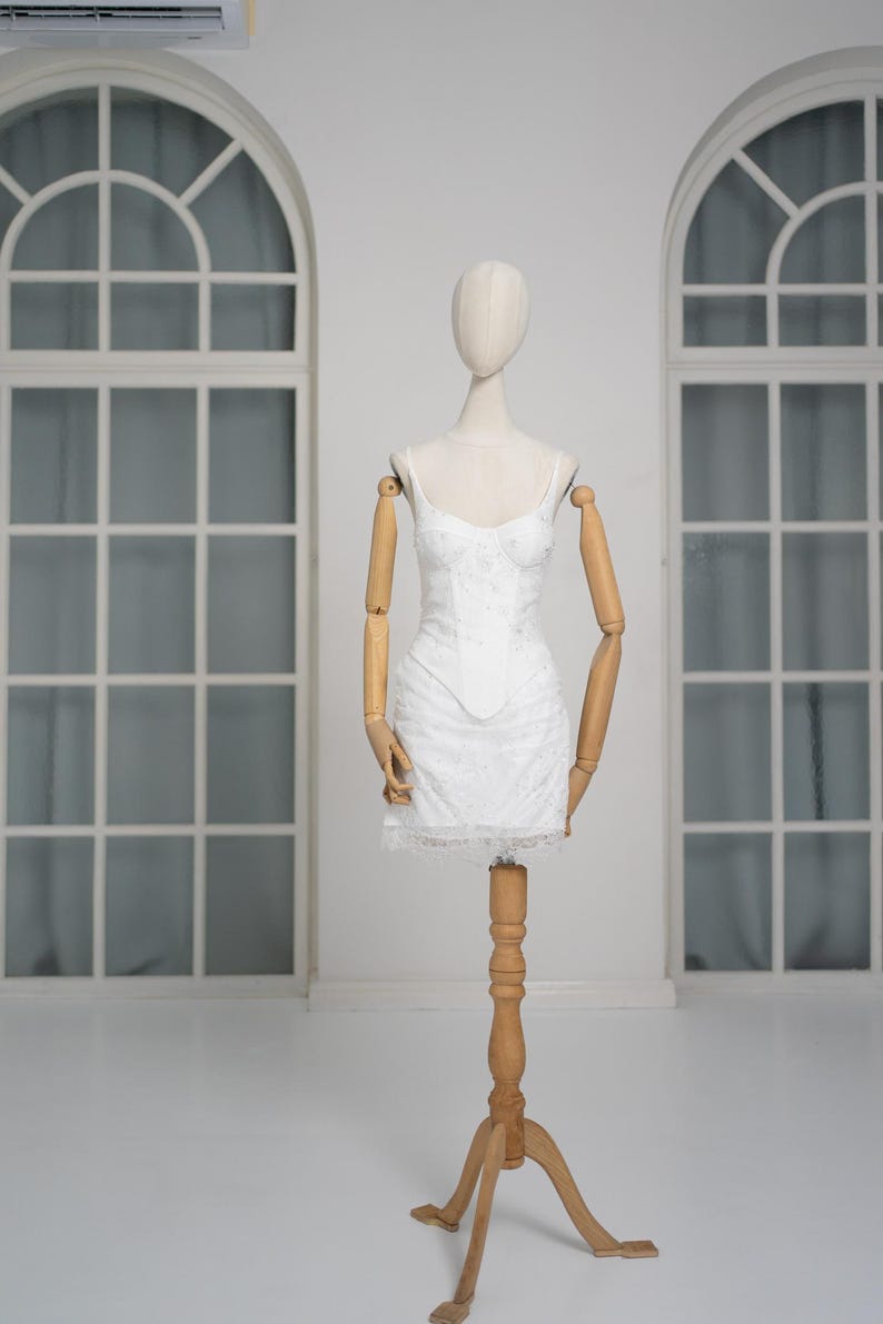 May include: A white, lace-detailed mini dress with spaghetti straps is displayed on a wooden mannequin. The dress features a fitted bodice and a flared skirt, with a delicate lace trim at the hem. The mannequin has articulated wooden arms.
