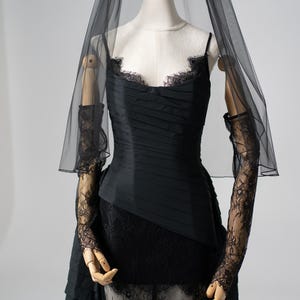 Op de afbeelding: Een zwarte jurk met een geplooid lijfje, spaghettibandjes en een met kant afgezette halslijn. De jurk heeft een gelaagde rok en is versierd met doorschijnende zwarte kanten handschoenen en een zwarte sluier. De jurk wordt op een mannequin getoond.