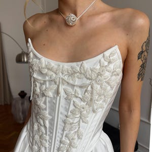 Puede incluir: Un top tipo corsé blanco sin tirantes con intrincados abalorios blancos en forma de hojas, acentuado con lentejuelas plateadas y delicados adornos de perlas. El corpiño está estructurado con costuras verticales y se combina con un collar de colgante esférico.