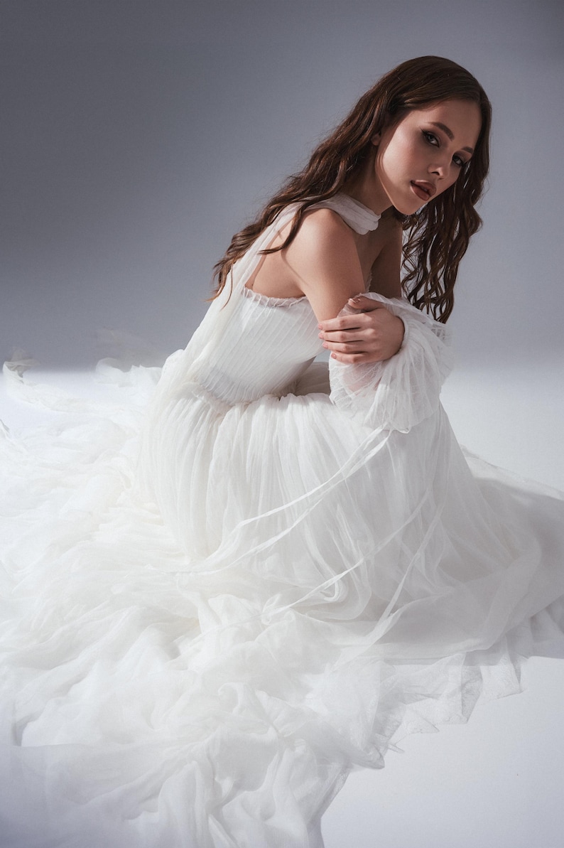May include: A white floor-length gown featuring a fitted bodice with vertical pleats, a high neckline, and long, voluminous sheer sleeves. The skirt is constructed from multiple layers of lightweight, flowing tulle fabric that pools on the floor.