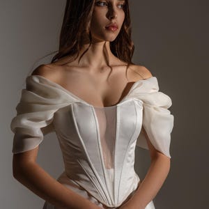 May include: A white off-the-shoulder wedding dress with a sheer neckline and structured bodice. The dress features a flowing skirt and sheer sleeves. The model is wearing the dress in a studio setting.