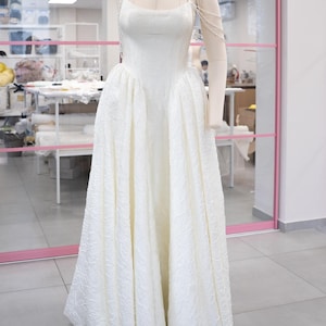 Può includere: Abito da sposa bianco su manichino. L'abito presenta un corpetto aderente con spalline sottili e una gonna ampia. Il tessuto ha un motivo floreale strutturato. Una collana di perle è drappeggiata sulla spalla del manichino.