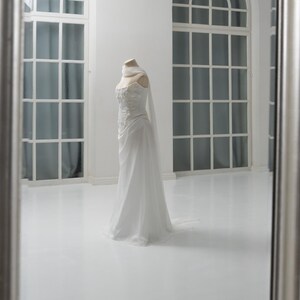 Puede incluir: Un vestido blanco largo hasta el suelo sobre un maniqu&iacute;, con un corpi&ntilde;o ajustado con intrincados bordados florales, un escote coraz&oacute;n y una falda fluida. Una bufanda de tela ligera y transparente est&aacute; colocada alrededor del cuello.