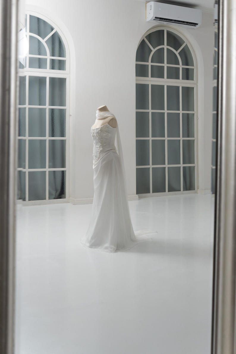 Puede incluir: Un vestido blanco largo hasta el suelo sobre un maniqu&iacute;, con un corpi&ntilde;o ajustado con intrincados bordados florales, un escote coraz&oacute;n y una falda fluida. Una bufanda de tela ligera y transparente est&aacute; colocada alrededor del cuello.