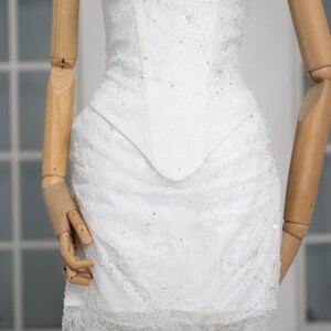 May include: White, embellished mini dress with a structured bodice and a lace-trimmed skirt. The dress features delicate beadwork and floral lace details, creating a romantic and elegant look. The dress is displayed on a wooden mannequin.