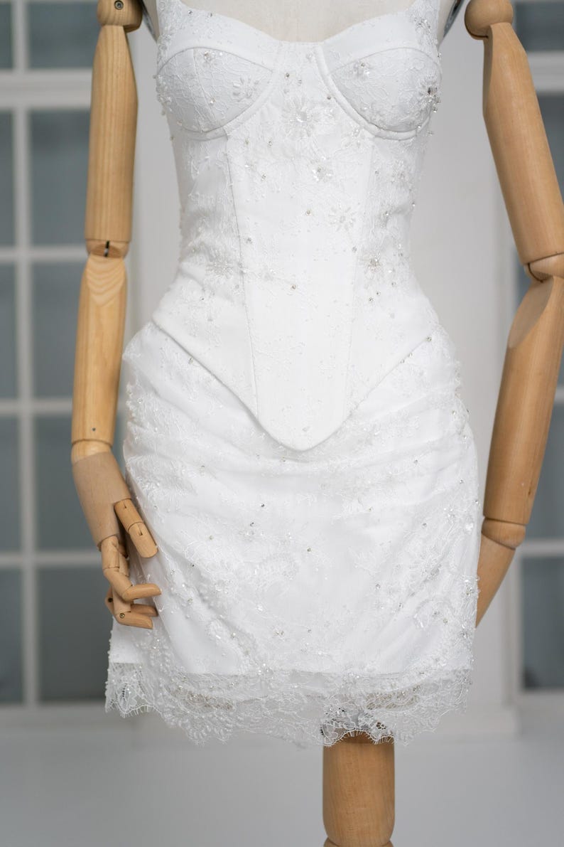 May include: White, embellished mini dress with a structured bodice and a lace-trimmed skirt. The dress features delicate beadwork and floral lace details, creating a romantic and elegant look. The dress is displayed on a wooden mannequin.