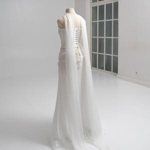 Puede incluir: Un vestido blanco largo hasta el suelo sobre un maniqu&iacute;, con una espalda estilo cors&eacute; con cierre de cordones. El vestido incluye paneles de tela transparente y fluida drapeados desde los hombros que se extienden hacia atr&aacute;s formando una larga cola.