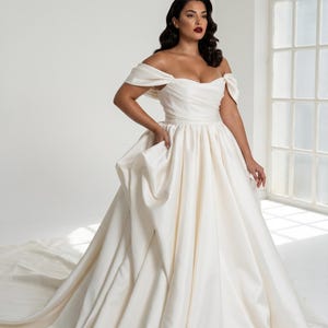 A woman wearing a long, off-the-shoulder white wedding dress stands in a bright, white room with a large window.