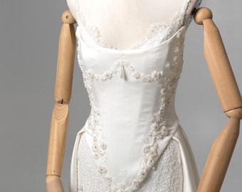 May include: A white wedding dress on a mannequin. The dress features a fitted bodice with floral and beaded embellishments, a high neckline, and a lace skirt. The dress is displayed on a wooden mannequin with articulated arms.