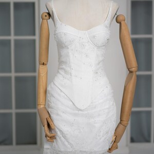 May include: A white, embellished mini dress with spaghetti straps and a fitted bodice. The dress features delicate beadwork and lace detailing, displayed on a mannequin with wooden arms and a neutral background. The dress is a bridal or formal wear item.