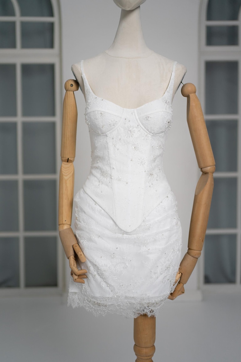 May include: A white, embellished mini dress with spaghetti straps and a fitted bodice. The dress features delicate beadwork and lace detailing, displayed on a mannequin with wooden arms and a neutral background. The dress is a bridal or formal wear item.