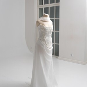 Puede incluir: Un vestido blanco sin tirantes sobre un maniqu&iacute; que presenta un corpi&ntilde;o ajustado con intrincados bordados florales y pedrer&iacute;a. La falda est&aacute; confeccionada con una tela fluida y transparente que cae hasta el suelo, con un pa&ntilde;uelo a juego al cuello.