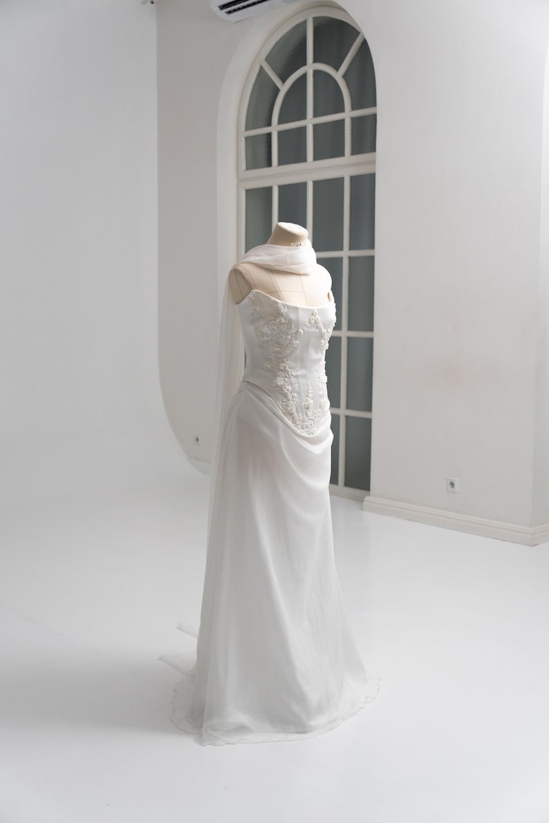Puede incluir: Un vestido blanco sin tirantes sobre un maniqu&iacute; que presenta un corpi&ntilde;o ajustado con intrincados bordados florales y pedrer&iacute;a. La falda est&aacute; confeccionada con una tela fluida y transparente que cae hasta el suelo, con un pa&ntilde;uelo a juego al cuello.