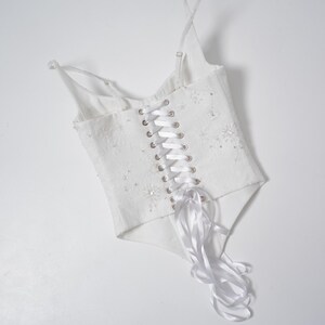 May include: White corset top with delicate spaghetti straps and a lace-up back. The bodice is embellished with subtle floral details and sparkling accents. A satin ribbon weaves through the eyelets, adding a touch of elegance to this garment.
