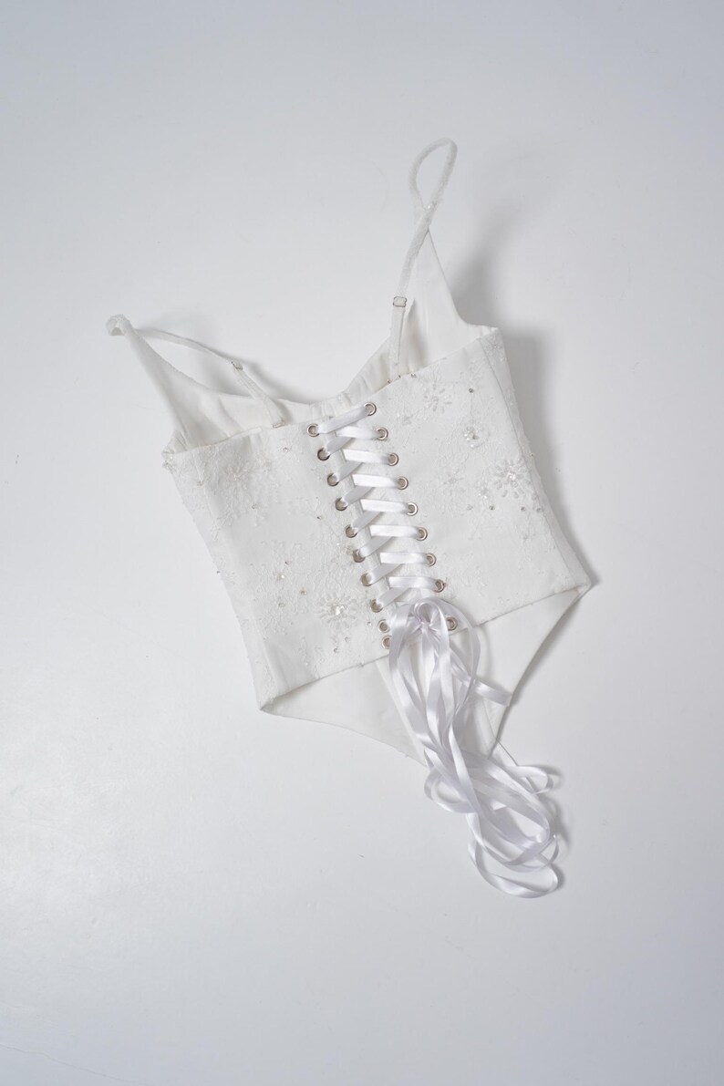 May include: White corset top with delicate spaghetti straps and a lace-up back. The bodice is embellished with subtle floral details and sparkling accents. A satin ribbon weaves through the eyelets, adding a touch of elegance to this garment.