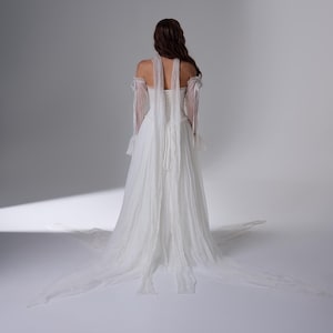 May include: A floor-length white gown featuring a corset-style bodice with lace-up back detailing, off-the-shoulder sheer sleeves with ruffled cuffs, and long flowing fabric panels extending from the shoulders down to a train on the floor.
