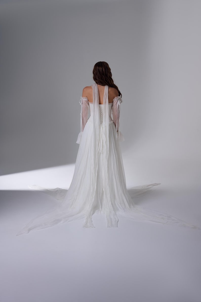 May include: A floor-length white gown featuring a corset-style bodice with lace-up back detailing, off-the-shoulder sheer sleeves with ruffled cuffs, and long flowing fabric panels extending from the shoulders down to a train on the floor.