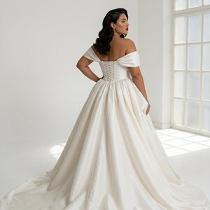 A woman wearing a long, off-the-shoulder white wedding dress with a full skirt and a train, standing in a studio with a large window in the background.
