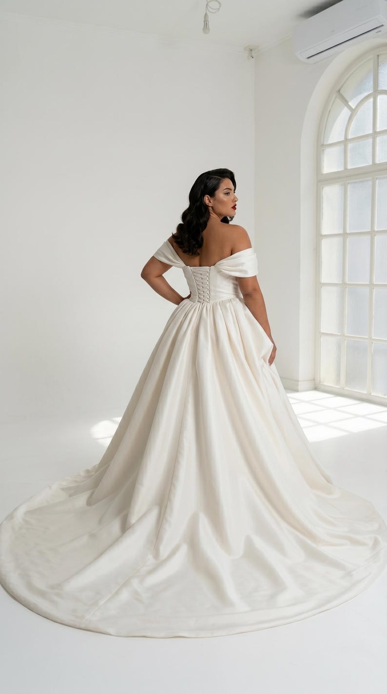 A woman wearing a long, off-the-shoulder white wedding dress with a full skirt and a train, standing in a studio with a large window in the background.