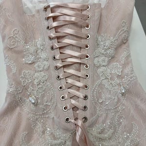 Pu&ograve; includere: Un top stile corsetto rosa pallido con sovrapposizione in pizzo, una chiusura centrale verticale con lacci in nastro di raso attraverso occhielli metallici color argento e ricami floreali bianchi con piccole perline e ciondoli perlati.