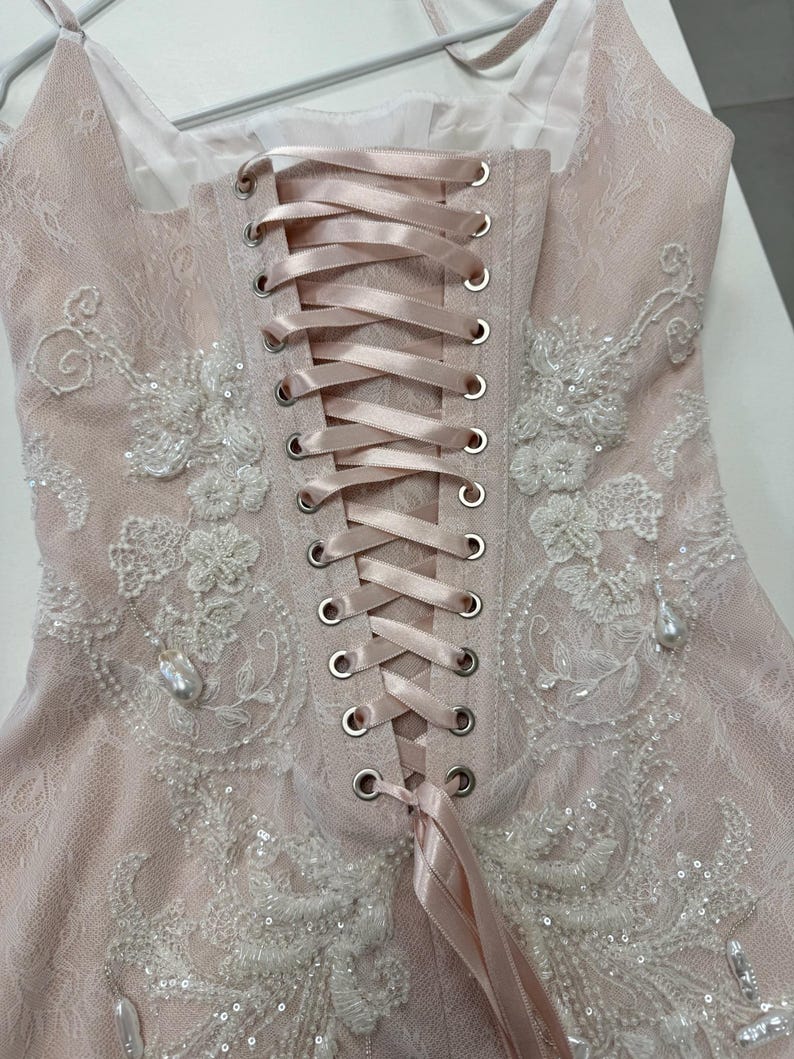 Pu&ograve; includere: Un top stile corsetto rosa pallido con sovrapposizione in pizzo, una chiusura centrale verticale con lacci in nastro di raso attraverso occhielli metallici color argento e ricami floreali bianchi con piccole perline e ciondoli perlati.