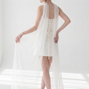 A woman in a white dress with a long train stands in a studio, her back to the camera, with her hair styled in an updo.