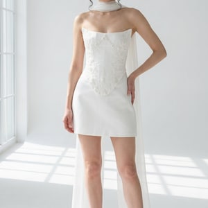 A woman wearing a white strapless dress with a long train, posing in a bright, white room with a window.