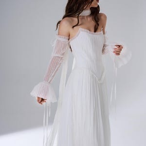 May include: A white floor-length dress featuring a pleated bodice and skirt, a straight neckline, and detachable sheer long sleeves with ruffled cuffs. The garment includes a matching sheer neck scarf and delicate ribbon ties at the shoulders.