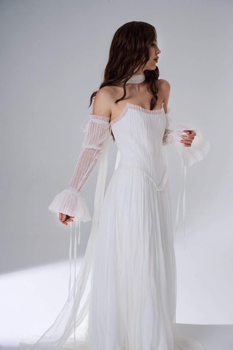 May include: A white floor-length dress featuring a pleated bodice and skirt, a straight neckline, and detachable sheer long sleeves with ruffled cuffs. The garment includes a matching sheer neck scarf and delicate ribbon ties at the shoulders.