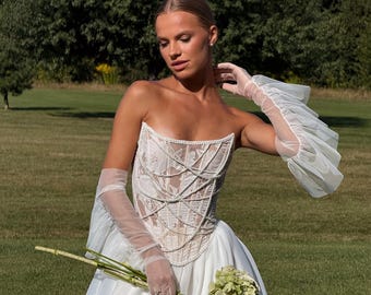Pode incluir: Vestido de noiva branco sem alças com corpete detalhado e saia ampla. O corpete apresenta rendas intrincadas e detalhes de pérolas. A noiva usa luvas longas e transparentes e segura um buquê de flores verdes e marrons. Fundo verde.