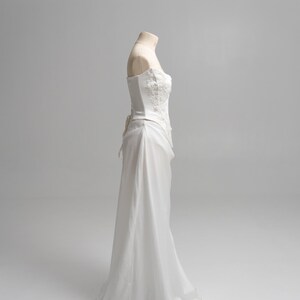 Puede incluir: Un vestido blanco sin tirantes con un corpi&ntilde;o tipo cors&eacute; estructurado con intrincados bordados florales y pedrer&iacute;a. La falda est&aacute; confeccionada con una tela transparente y fluida que cae desde la cintura hasta el suelo, creando una silueta suave.