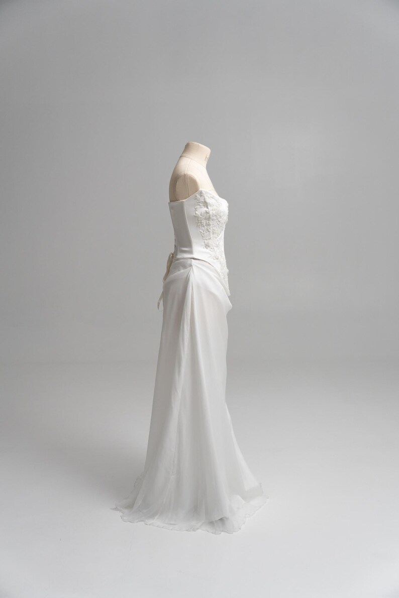 Puede incluir: Un vestido blanco sin tirantes con un corpi&ntilde;o tipo cors&eacute; estructurado con intrincados bordados florales y pedrer&iacute;a. La falda est&aacute; confeccionada con una tela transparente y fluida que cae desde la cintura hasta el suelo, creando una silueta suave.