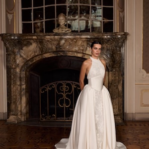 May include: A floor-length ivory gown featuring a high-neck lace bodice with a structured satin corset waist and a voluminous satin overskirt that opens to reveal a lace-detailed column skirt underneath, creating a layered silhouette.